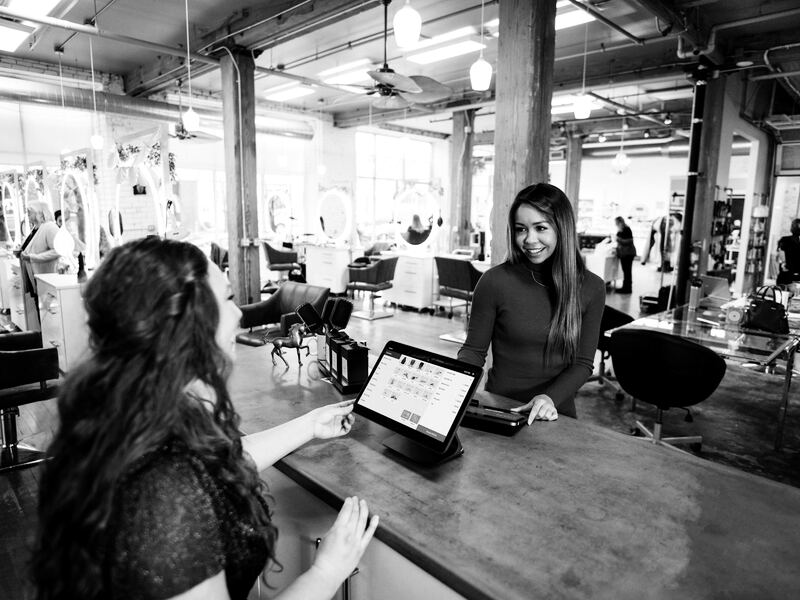 Two women chatting across a shop counter while one shows the other details on a tablet